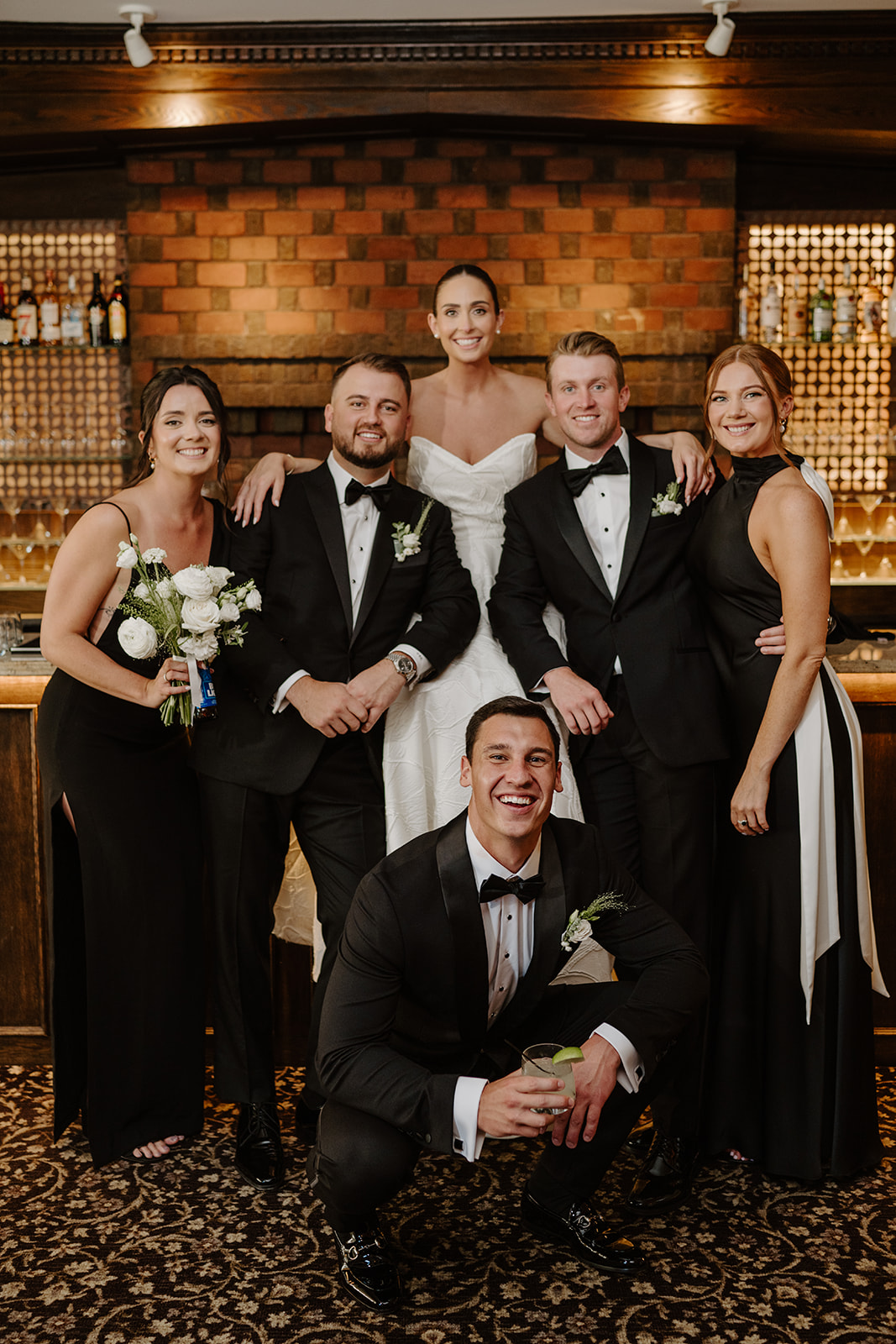 Groom crouching with a drink in front of his bride and wedding party members posing at the bar.